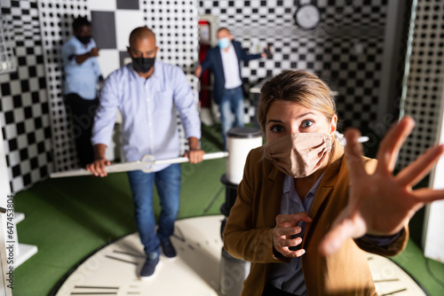 Young woman with friends wearing face masks for viral protection solving puzzles in quest room, reaching for next key