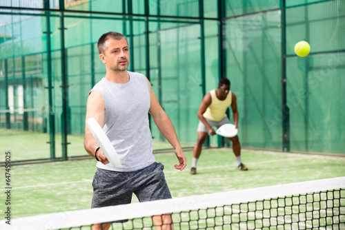 Young adult man playing paddle tennis doubles match with male partner at warm sunny day © JackF