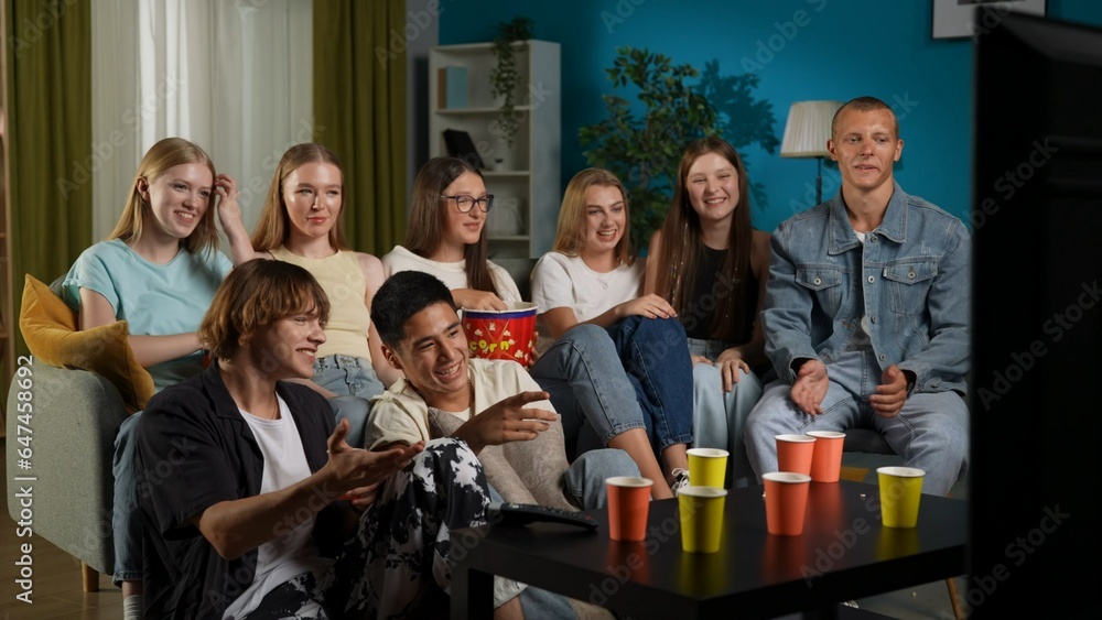 Group of teens, young people, friends sitting on a couch, partying, watching TV, movie, show. They are eating popcorn, laughing and chatting, joking around. - obrazy, fototapety, plakaty 