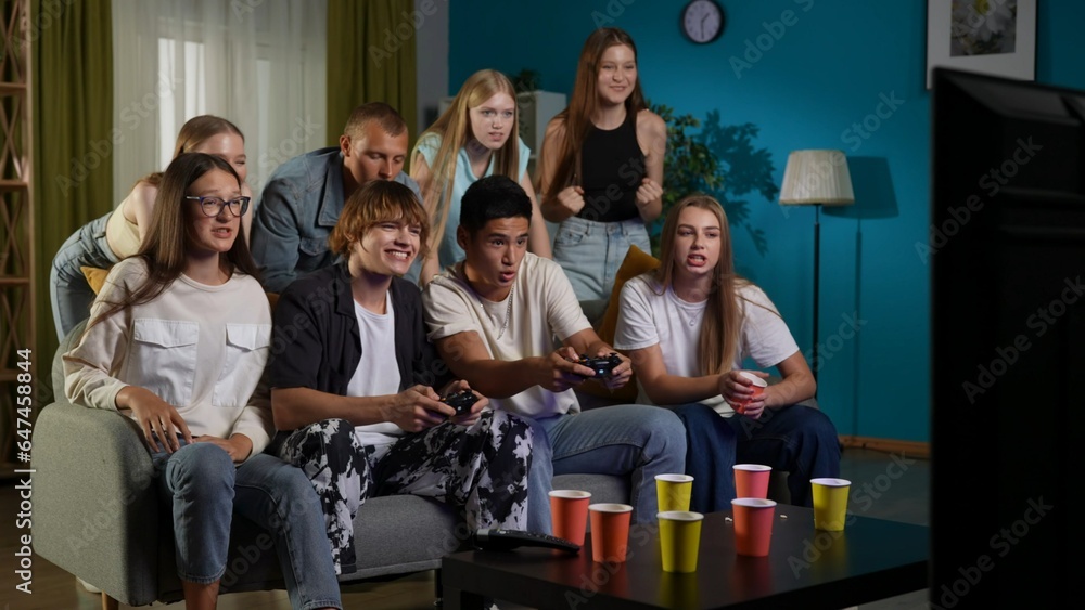 Group of teens, young people, friends sitting and standing around the TV and cheering, encouraging two guys, boys who are playing a console, gaming, play-fighting. Full-size.