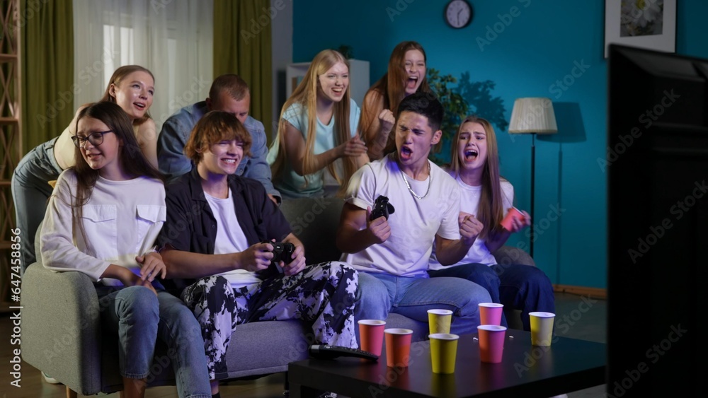Group of teens, young people, friends sitting and standing around two guys, boys who were playing a console, gaming. One of them is celebrating his win emotionally. Full-size.