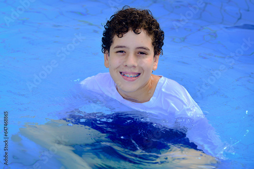 man in swimming pool, child in swimming pool, child in pool, boy in pool ,person in pool, white boy, curly hair, smiling, happy boy, with orthodontic appliance, beachwear photo