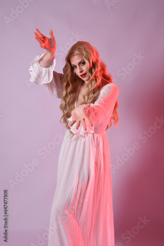 Close up portrait of beautiful blonde model wearing elegant white halloween gown. arms reaching out like hungry vampire. isolated on studio background with red cinematic moody lighting.