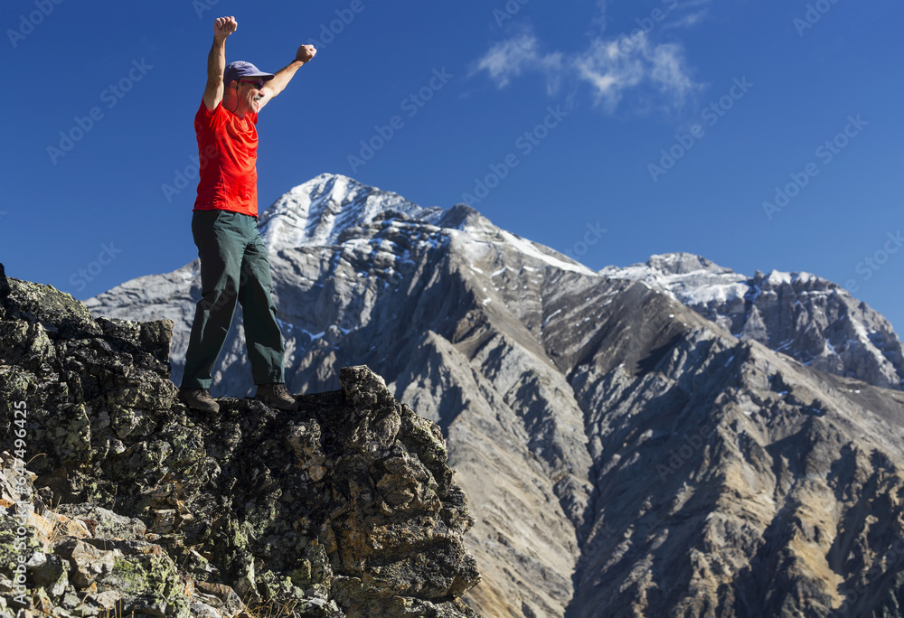 Male Hiker Standing With Arms Raised In The Air On Top Of Mountain Ridge Overlooking Snow Peaked Mountains With Blue Sky And Clouds, Kananaskis Provincial Park; Alberta, Canada Male Hiker Standing With Arms Raised In The Air On Top Of Mountain Ridge Overlooking Snow Peaked Mountains With Blue Sky And Clouds, Kananaskis Provincial Park; Alberta, Canada