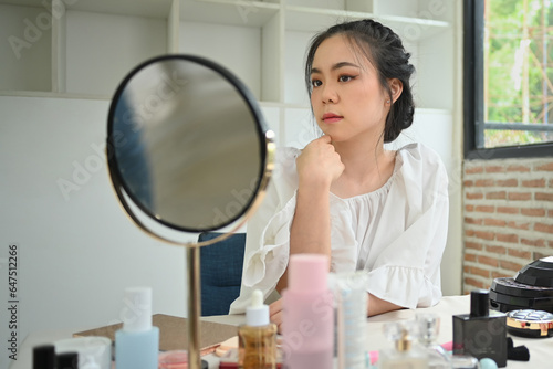 Charming young asian woman sitting in front of mirror and looking at reflection. Beauty, cosmetic and makeup concept