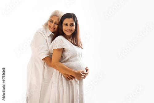 A PREGNANT YOUNG WOMAN STANDING AND SMILING WITH MOTHER EMBRACING HER BABY BUMP photo