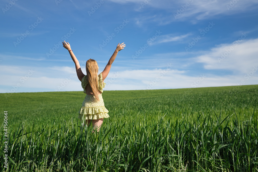 Young, beautiful, blonde woman in a yellow dress, arms raised in the middle of a green wheat field, seen from the back, alone, calm and peaceful. Concept relaxation, tranquility, calm, fields, meadows Young, beautiful, blonde woman in a yellow dress, arms raised in the middle of a green wheat field, seen from the back, alone, calm and peaceful. Concept relaxation, tranquility, calm, fields, meadows