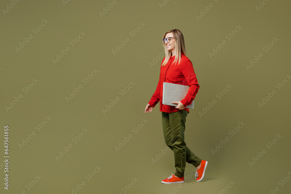 Full body side profile view young IT woman she wearing red shirt casual clothes glasses holding closed laptop pc computer look aside isolated on plain pastel green background studio Lifestyle concept. Full body side profile view young IT woman she wearing red shirt casual clothes glasses holding closed laptop pc computer look aside isolated on plain pastel green background studio Lifestyle concept.