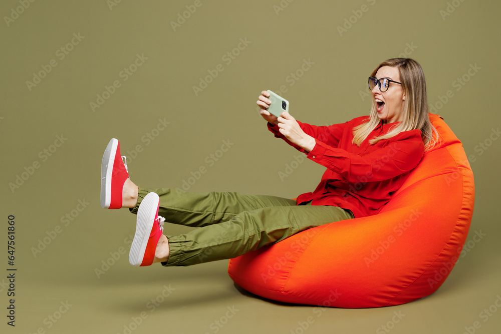 Full body young woman in red shirt casual clothes glasses sit in bag chair use play racing app on mobile cell phone hold gadget smartphone for pc video game isolated on plain pastel green background Full body young woman in red shirt casual clothes glasses sit in bag chair use play racing app on mobile cell phone hold gadget smartphone for pc video game isolated on plain pastel green background
