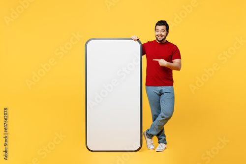 Full body young Indian man wears red t-shirt casual clothes point finger on big huge blank screen mobile cell phone smartphone with area isolated on plain yellow orange background. Lifestyle concept.