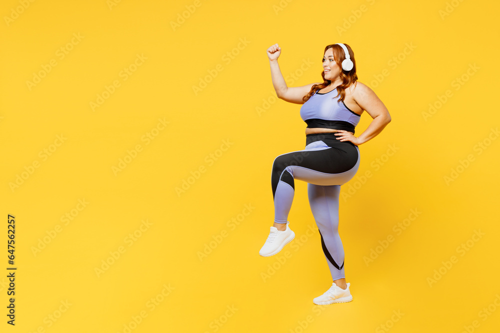 Full body young plus size big fat fit woman wear blue top warm up training listen to music in headphones raise up leg hand isolated on plain yellow background studio home gym. Workout sport concept.