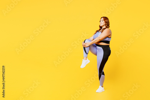 Full body side view young chubby plus size big fat fit woman wear blue top warm up training raise up leg do stretch exercises isolated on plain yellow background studio home gym Workout sport concept
