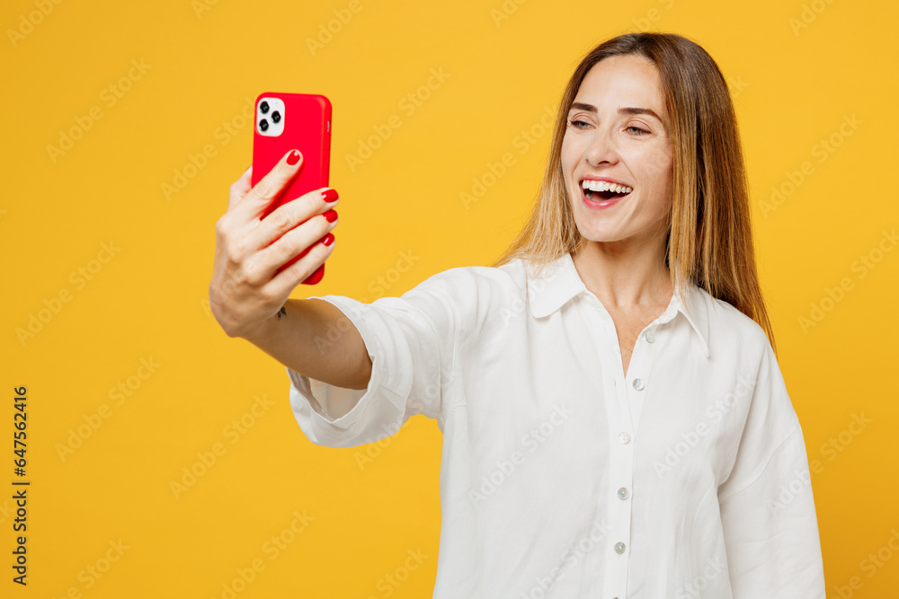 Young caucasian happy woman wears white shirt casual clothes doing selfie shot on mobile cell phone post photo on social network isolated on plain yellow background studio portrait. Lifestyle concept. Young caucasian happy woman wears white shirt casual clothes doing selfie shot on mobile cell phone post photo on social network isolated on plain yellow background studio portrait. Lifestyle concept.