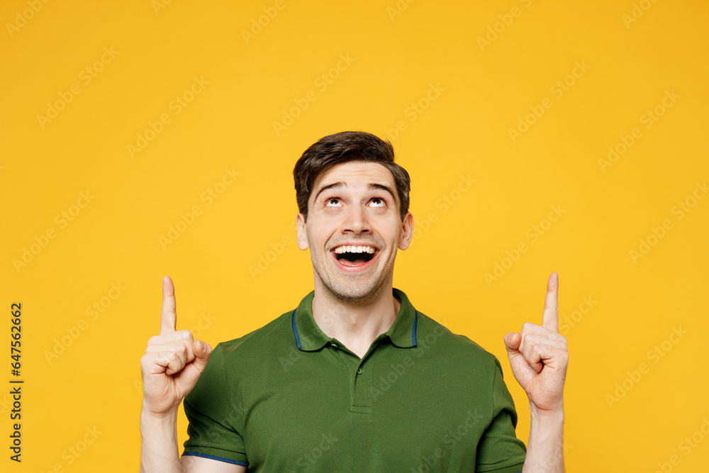 Young smiling man wears green t-shirt casual clothes point index finger overhead indicate on workspace area copy space mock up isolated on plain yellow background studio portrait. Lifestyle concept. Young smiling man wears green t-shirt casual clothes point index finger overhead indicate on workspace area copy space mock up isolated on plain yellow background studio portrait. Lifestyle concept.