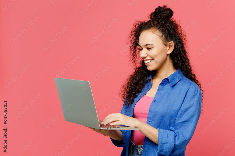 Side view young happy IT woman of African American ethnicity she wear blue shirt casual clothes hold use work on laptop pc computer isolated on plain pastel pink background studio. Lifestyle concept. Side view young happy IT woman of African American ethnicity she wear blue shirt casual clothes hold use work on laptop pc computer isolated on plain pastel pink background studio. Lifestyle concept.