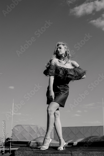 Woman wearing shoulderless dress adorned with ruffle, high heels shoes stand on roof over sky, holding hand on waist.
