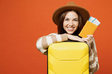 Traveler smiling woman wear brown sweater hat hold bag passport ticket isolated on plain orange red background. Tourist travel abroad in free spare time rest getaway. Air flight trip journey concept.