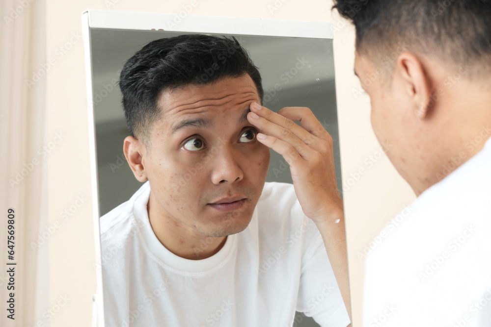 Obraz premium Photo of anxious Asian young man looking in the mirror worried about wrinkles or acne on unhealthy skin.