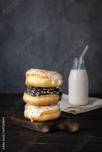 Donuts in white and chocolate glaze with multi-colored sprinkles on a wooden board. A bottle of milk in the background. Dark background. Space for text