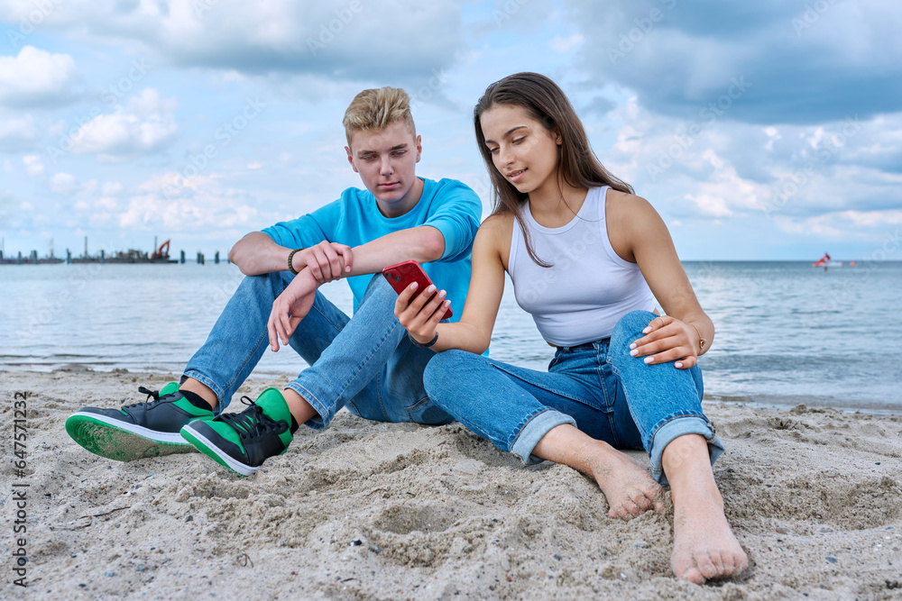 Teenage friends guy and girl sitting together on beach, talking looking at smartphone screen Teenage friends guy and girl sitting together on beach, talking looking at smartphone screen