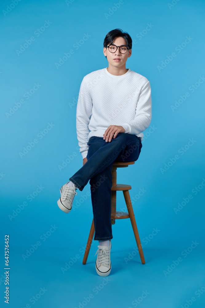 Vertical shot of a young cheerful man sitting on a stool isolated on blue background