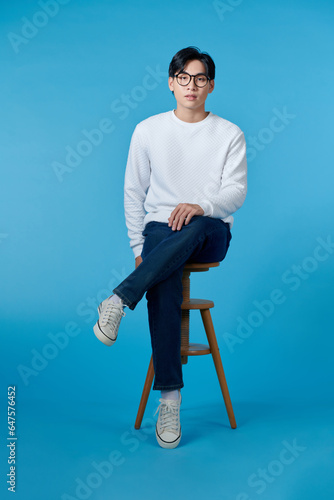 Obraz na płótnie Vertical shot of a young cheerful man sitting on a stool isolated on blue background w salonie
