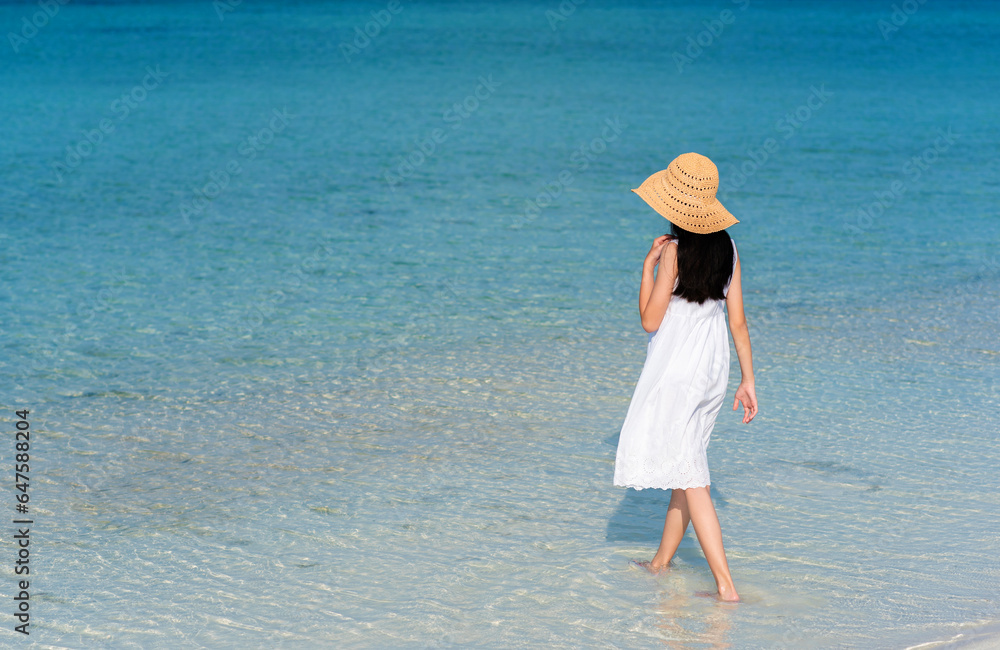 Back of young Asian girl walking on white sand beach with clear blue sea and sky. Teenager girl wearing sun straw hat, white dress enjoy vacation. Outdoor summer vacation travel concept, copy space.