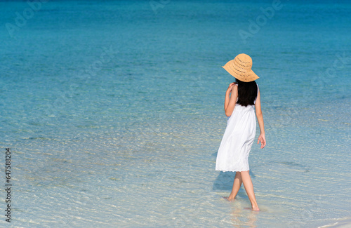 Back of young Asian girl walking on white sand beach with clear blue sea and sky. Teenager girl wearing sun straw hat, white dress enjoy vacation. Outdoor summer vacation travel concept, copy space.