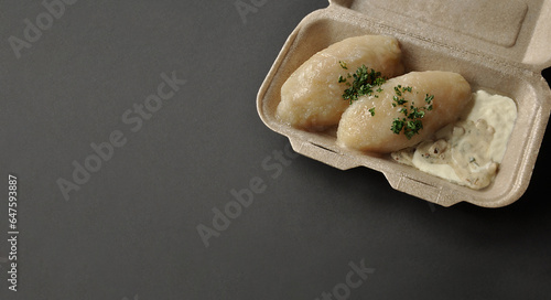 Lithuanian national dish - potato dumplings (zeppelins) with meat and mushroom sauce, sour cream, parsley. Takeaway box. Sustainable packaging. Dark background