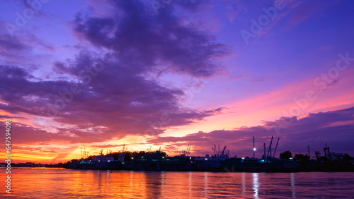 Beautiful Dramatic Sunset Sky over silhouette nautical Vessels moored along the river bank with colorful sunlight reflection on river surface in evening time