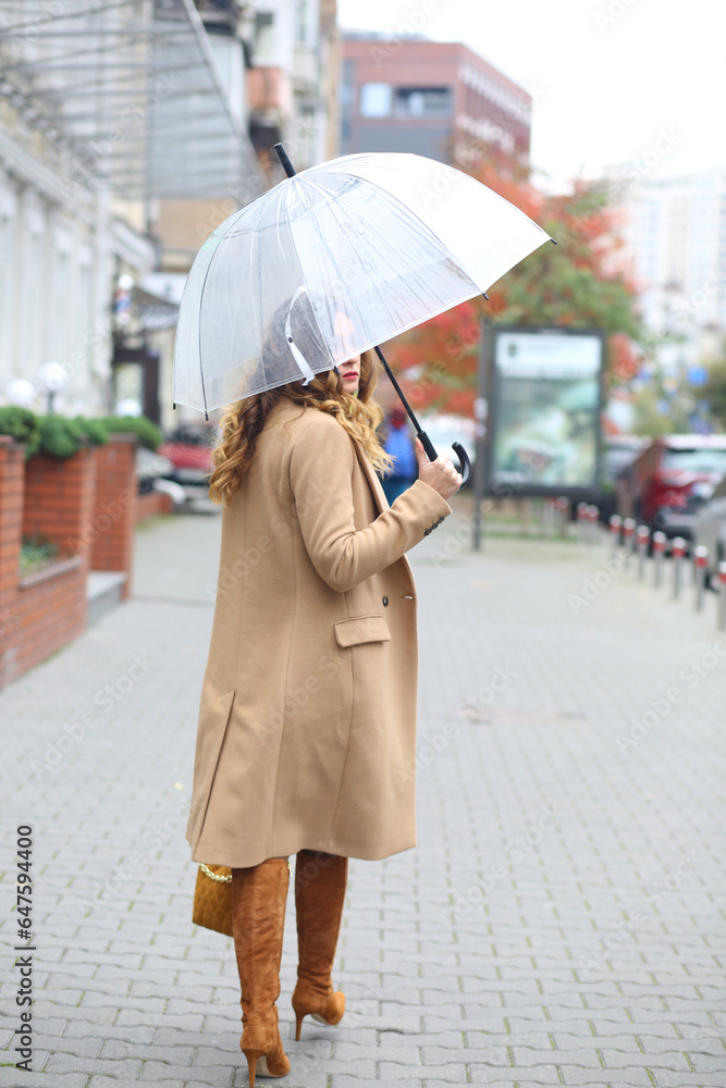 Naklejka premium A girl in a beige coat with a transparent umbrella walking down the street. Europe. Travels. autumn Coldly. Weather forecast.