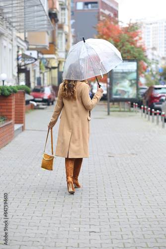 A girl in a beige coat with a transparent umbrella walking down the street. Europe. Travels. autumn Coldly. Weather forecast.