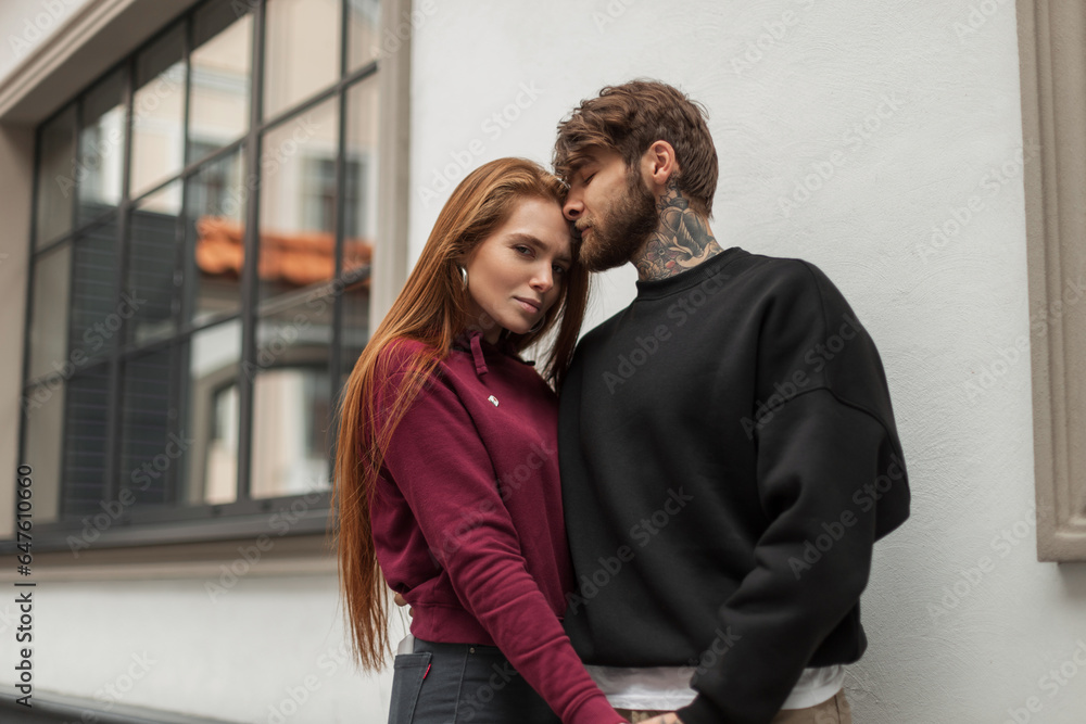 Beautiful fashion couple of lovers in stylish clothes with a pullover are standing on the street. Handsome hipster man in a black sweatshirt and a beautiful redhead woman in a red hoodie hug outdoors