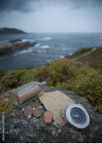 A traveler's compass, an old sheet of paper, a glass bottle with a message inside and several metal coins lie on a rock against the backdrop of a sea bay