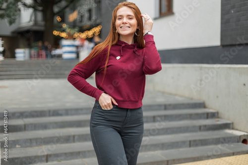 Cheerful beautiful fresh emotional woman with red hair with a smile in a fashionable red hoodie walks on the street