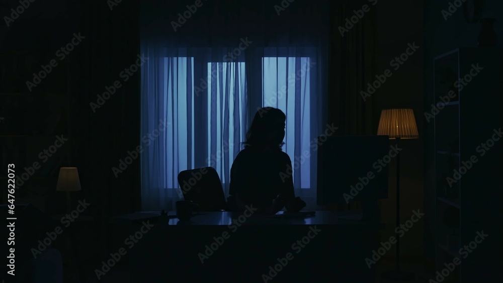 Everyday life creative concept. Woman in casual clothing sitting at the desk in dark room and works on the computer. Everyday life creative concept. Woman in casual clothing sitting at the desk in dark room and works on the computer.