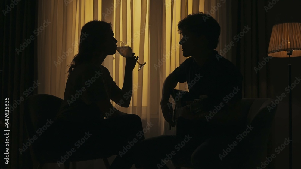 Everyday life creative concept. Young couple sitting in the room, man playing the guitar, woman drinks glass of wine.