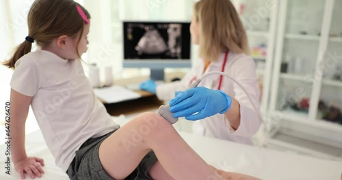 Professional doctor checks knee joint of schoolgirl with ultrasound machine. Attentive woman specialist in medical uniform works in clinic office