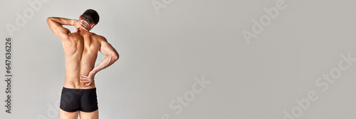 Back and neck pains. Muscular young man with relief body standing in underwear over grey studio background. Concept of men's health and beauty, body care, fitness, wellness. Banner. Copy space for ad