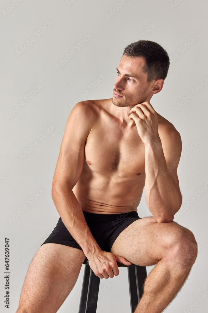 Studio portrait of handsome young man with really, muscular, beautiful body posing on chair in underwear against grey background. Concept of men's health and beauty, body care, fitness, wellness, ad