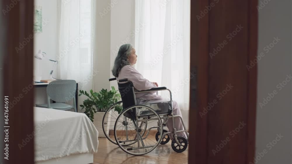 Long shot of depressed elderly woman with disability in wheelchair looking at window in nursing home Long shot of depressed elderly woman with disability in wheelchair looking at window in nursing home