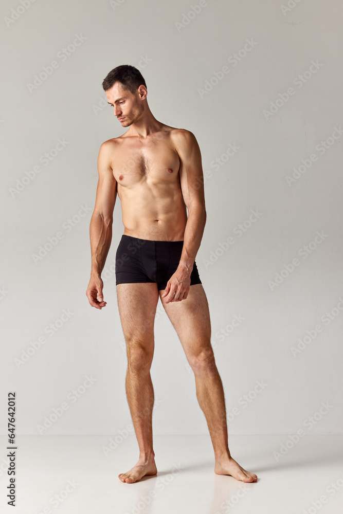 Full-length image of handsome young man with strong, muscular, relief body standing in underwear against grey studio background. Concept of men's health and beauty, body care, fitness, wellness, ad