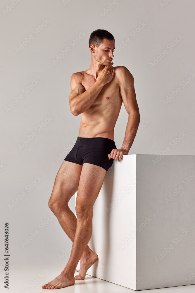 Full-length image of handsome, shirtless man with muscular body posing in underwear against grey studio background. Concept of men's health and beauty, body care, fitness, wellness, ad