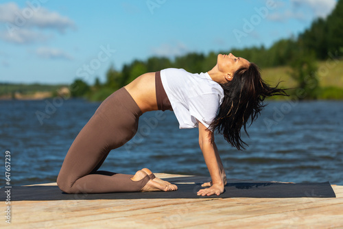 A woman does yoga, performs Ardha Chandrasana exercises, half-bridge pose, works out alone in sportswear on the shore of a lake on a sunny morning © Sergey Chayko