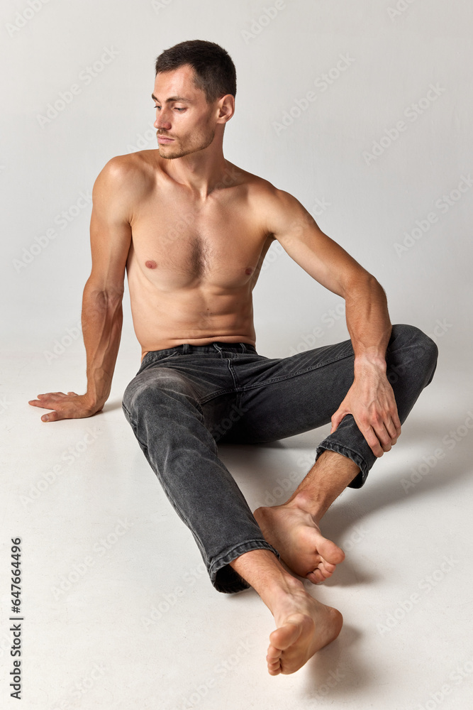 Obraz premium Portrait of handsome man in his 20s with muscular body shape posing shirtless in jeans against grey studio background. Concept of men's health and beauty, body care, fitness, wellness, ad