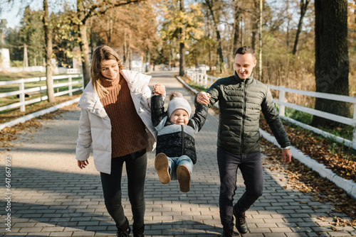 Mother, father throws up kid and running in street near forest in nature. Mom, dad hold hands son child walking in park at sunset. Family spending time together on vacation. Concept of autumn holiday.