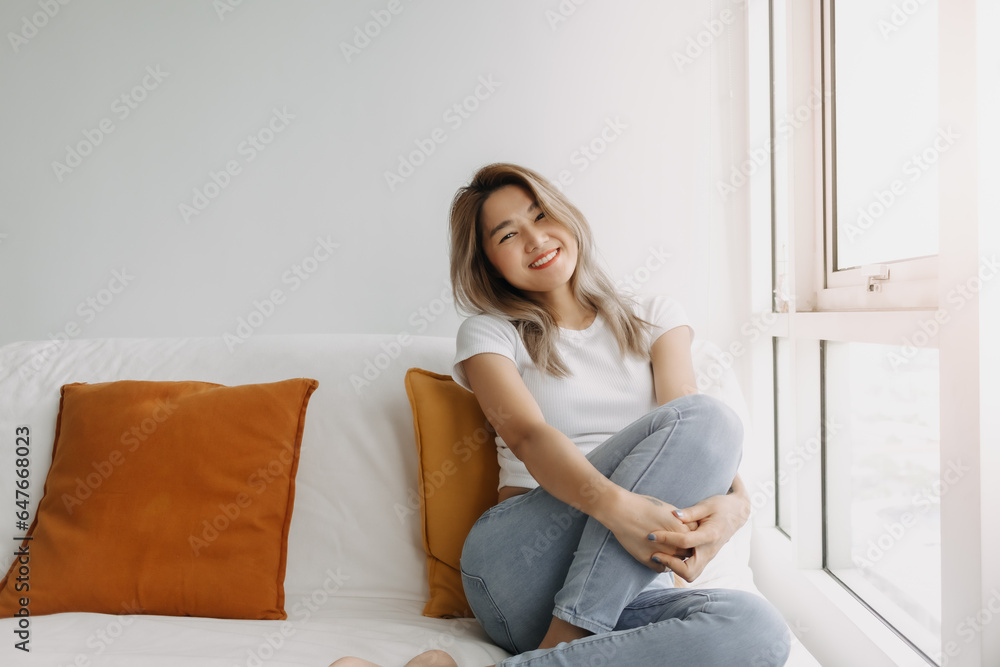 Happy Asian Thai women smiling and sitting on sofa at apartment room, looking at the camera with good life ralaxing.