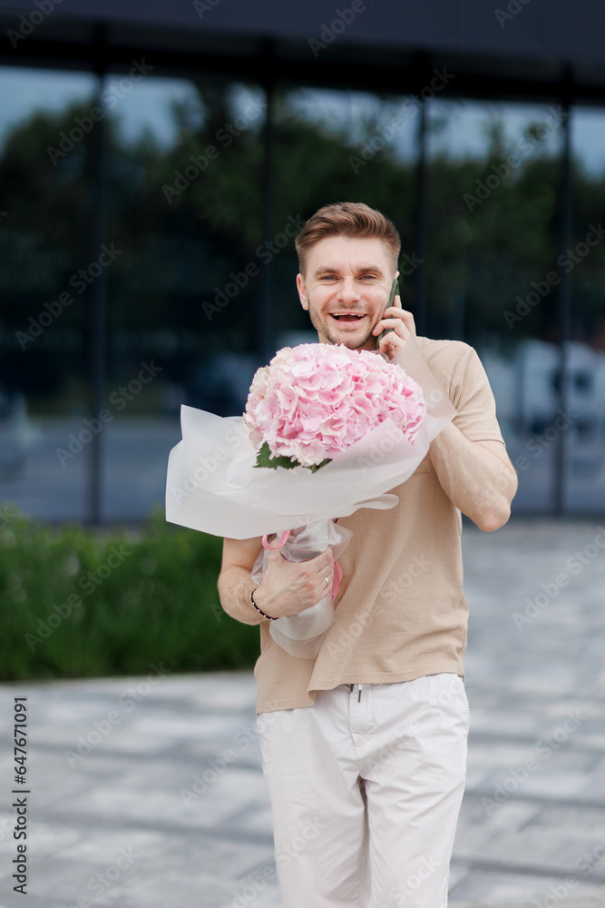 Fototapeta premium Flower delivery. Smiling delivery man with luxury bouquet of hydrangea flowers talks on smartphone with client on territory modern residential building. Gift for Valentines day, Birthday, Anniversary
