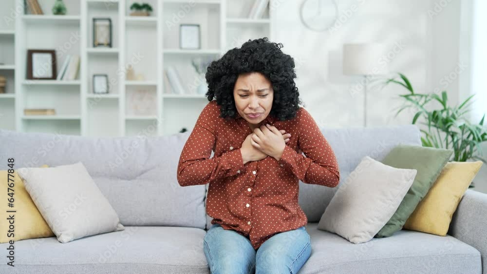 Sick young african american female suffering from chest tension, heart attack sitting on the sofa in living room at home. Black woman holding her chest with her hands, needs urgent medical attention Sick young african american female suffering from chest tension, heart attack sitting on the sofa in living room at home. Black woman holding her chest with her hands, needs urgent medical attention