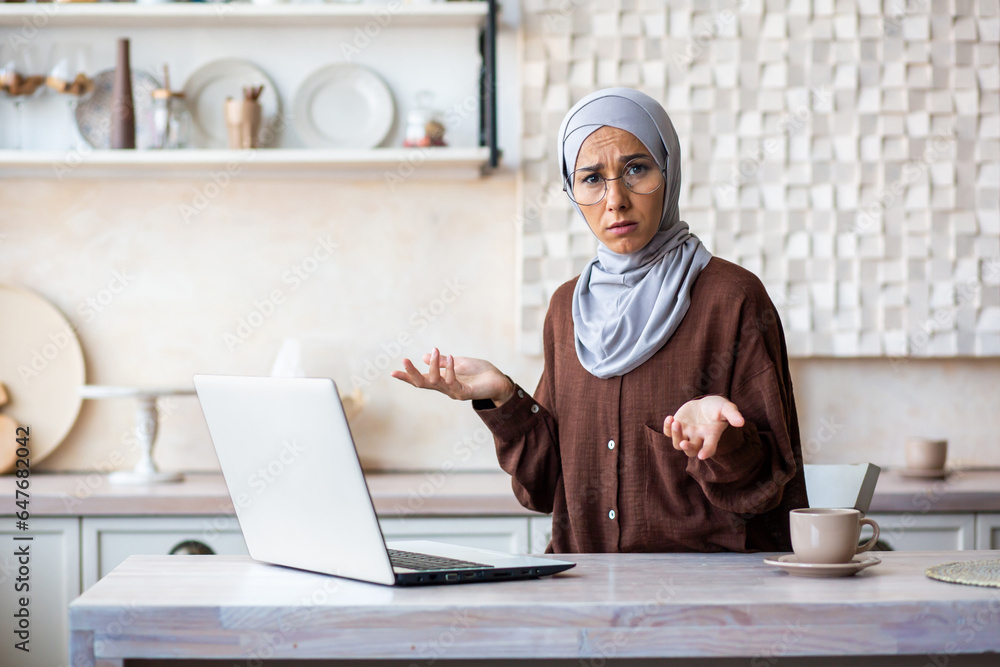 Portrait of a young Arab woman in hijab looking worriedly at the camera and spreading her hands. Sitting at home in the kitchen on a laptop. Portrait of a young Arab woman in hijab looking worriedly at the camera and spreading her hands. Sitting at home in the kitchen on a laptop.